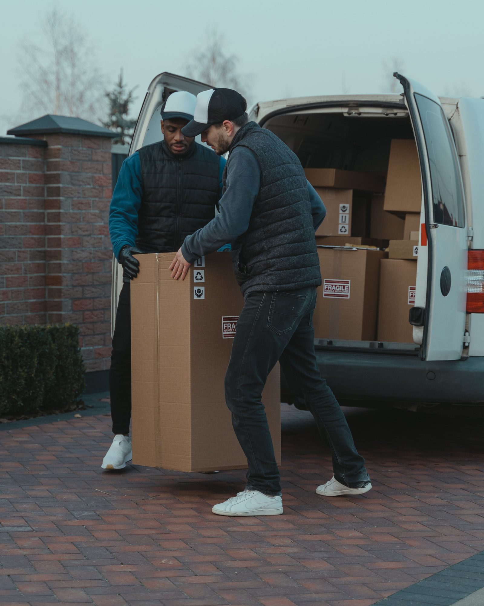 Two men lift a tall cardboard box out of the back of a white van.