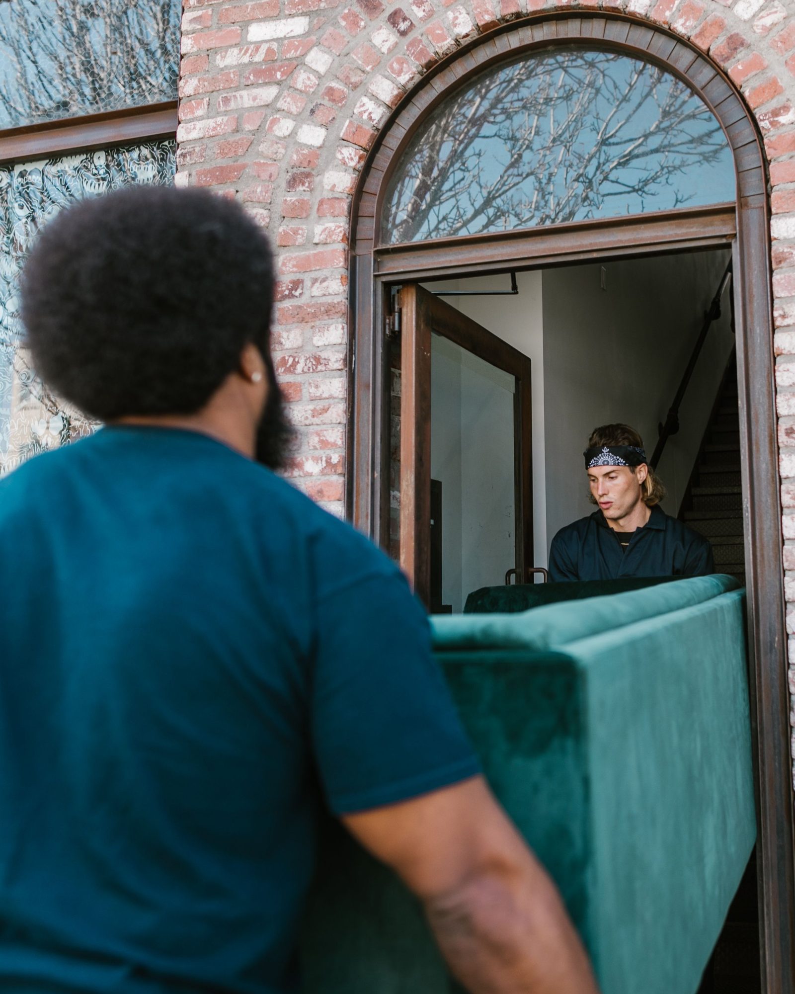 Two people carry a green sofa through an arched brick doorway.