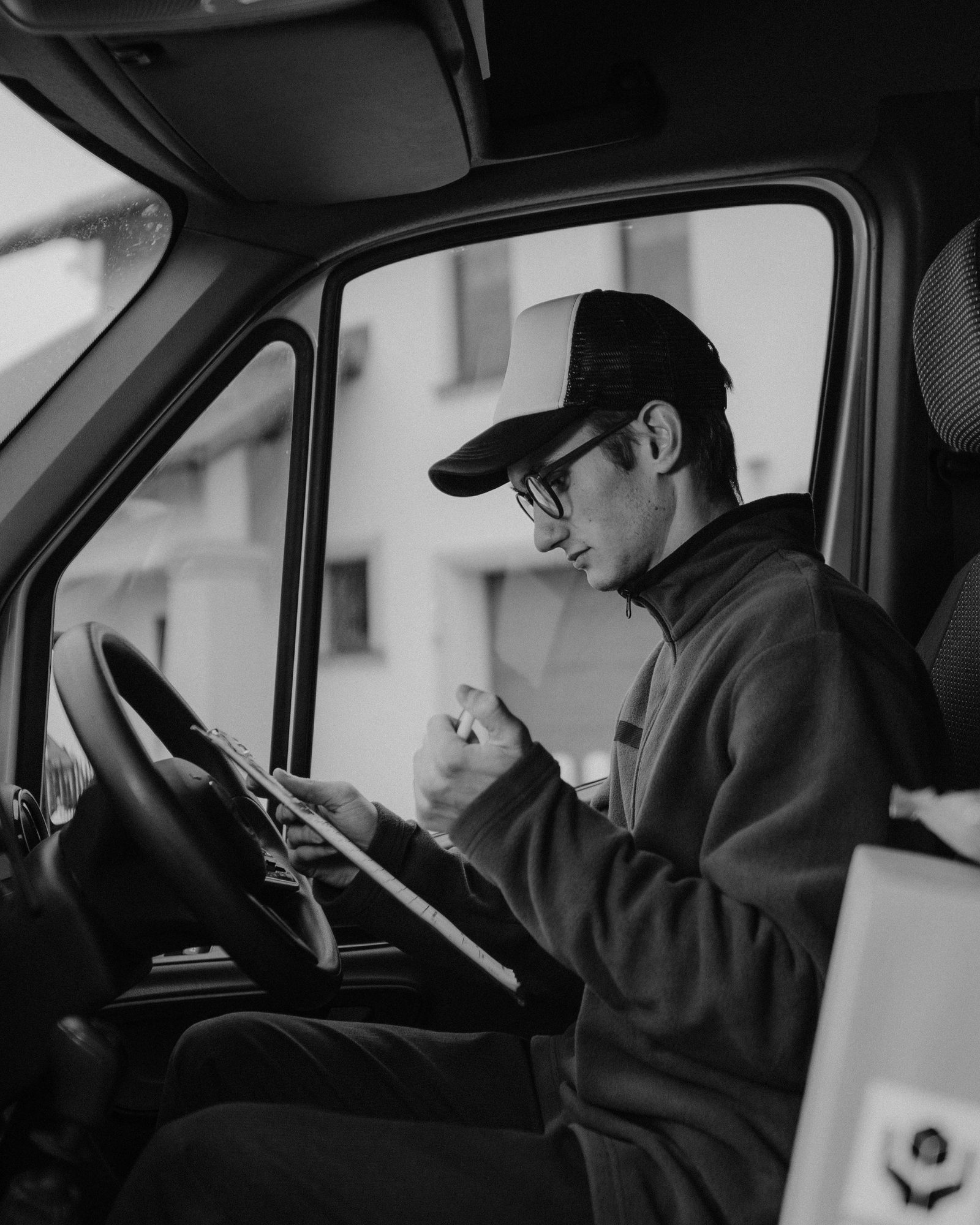 A man in a cap reads paperwork at the wheel of a van.